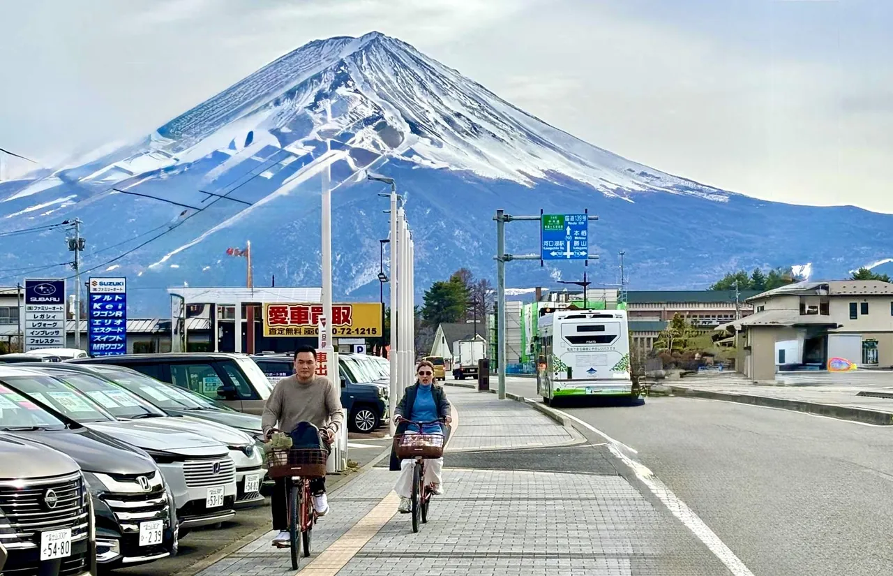 Two people ride bicycles on a paved sidewalk along a street with parked cars, with a large snow-capped mountain dominating the background.