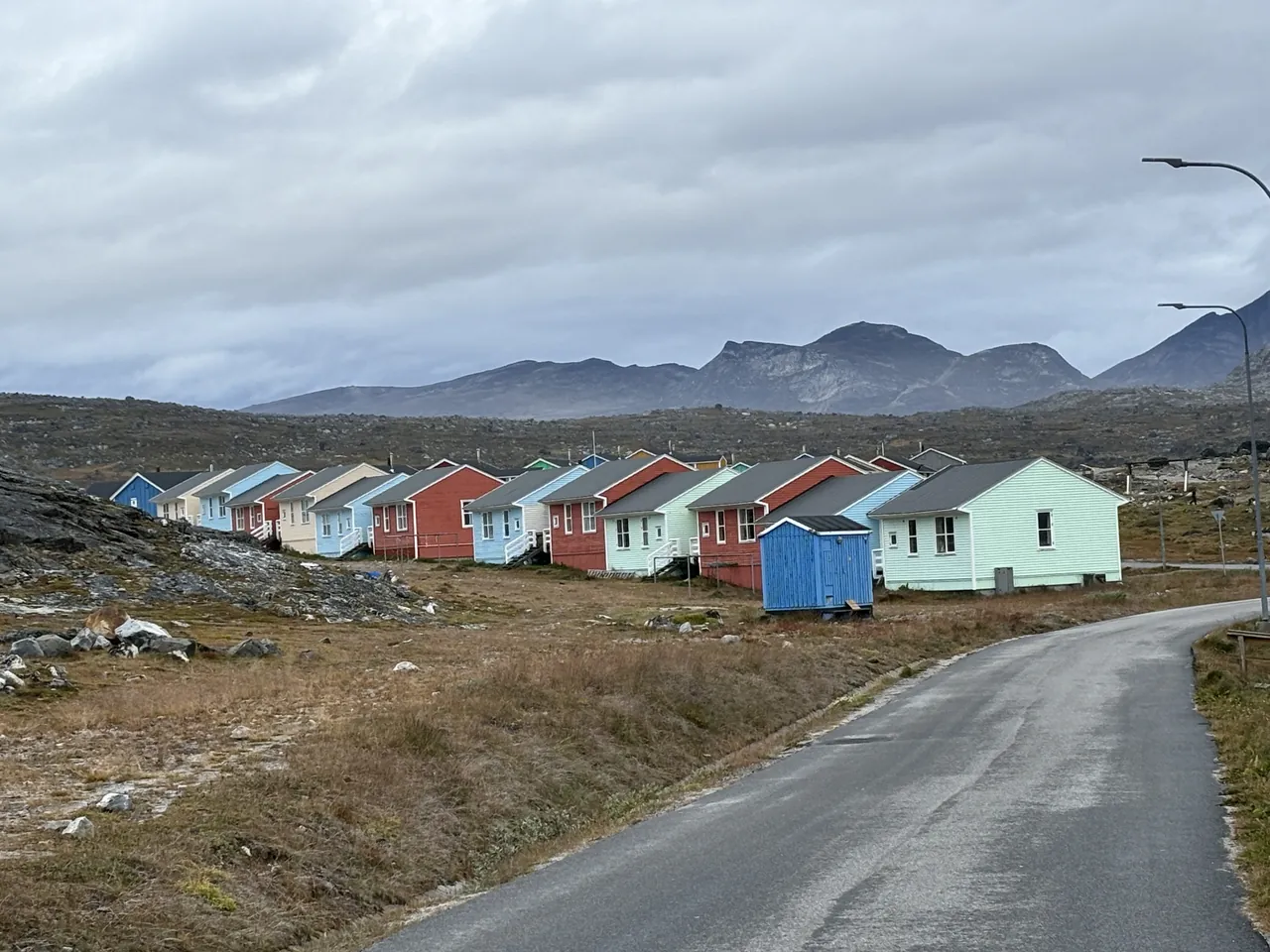 A row of brightly colored single-story houses lines a paved road beneath a cloudy sky, with barren hills and mountains in the background.