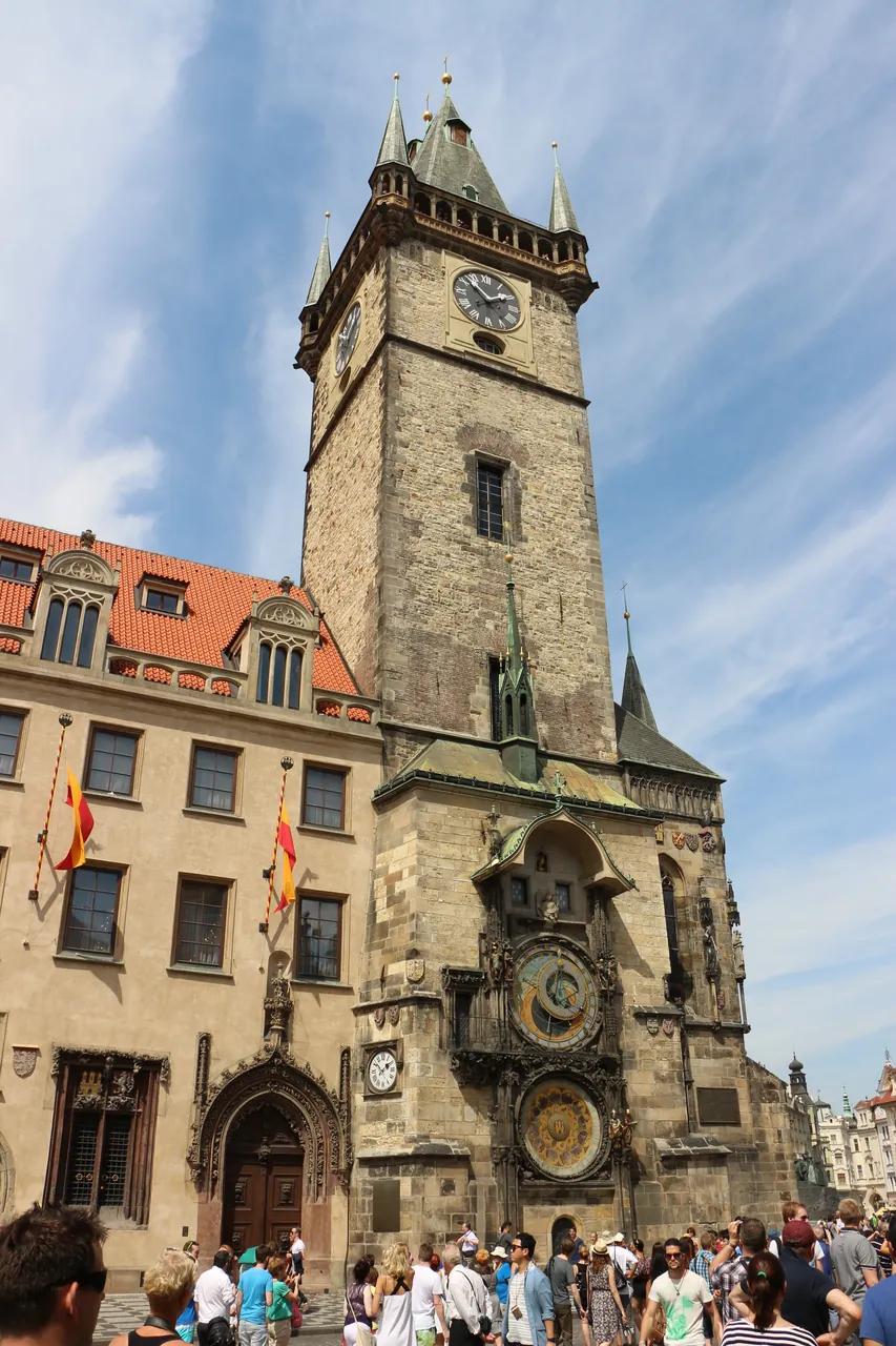 A full-frame upward shot shows the Prague Astronomical Clock tower with crowds of tourists gathered at the base under a partly cloudy sky.