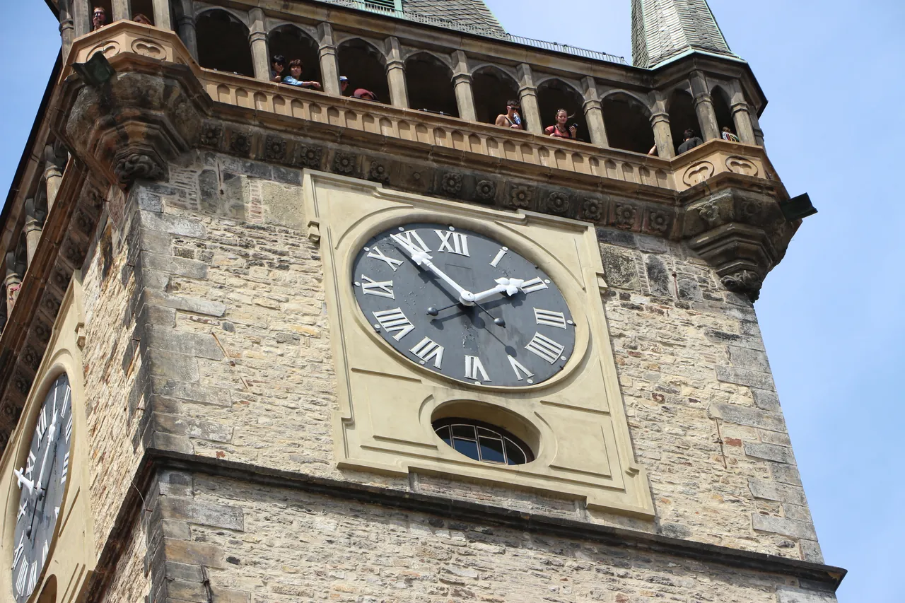 A close-up view of the stone exterior and clock face of a historic tower, with visitors visible on the upper viewing gallery.