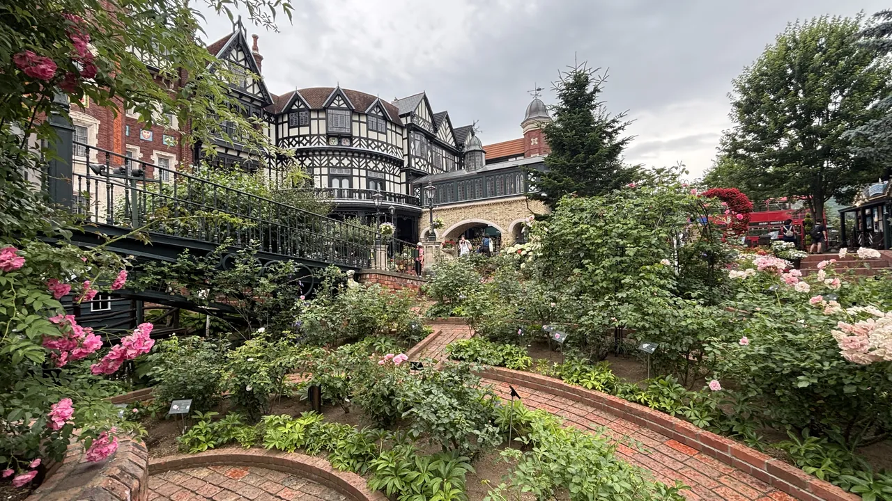 A rose garden with brick pathways is set in front of Tudor-style buildings under an overcast sky.